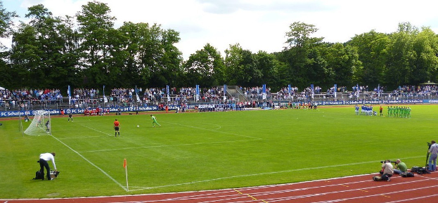 Eine Gruppe von Menschen, die Fußball auf einem Feld mit einem Tor spielen, umgeben von einer großen Menge, Fahnen, Bäumen und einem bewölkten Himmel.