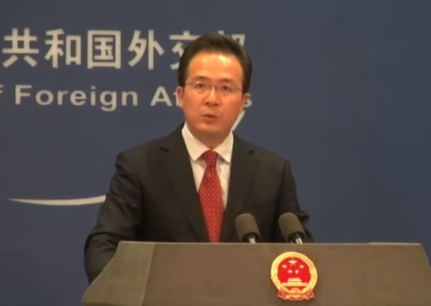 Ein Mann im Anzug und Krawatte steht an einem Podium mit zwei Mikrofonen und spricht als ausländischer Agent, mit einem Logo auf dem Podium und einem Banner mit Text dahinter.