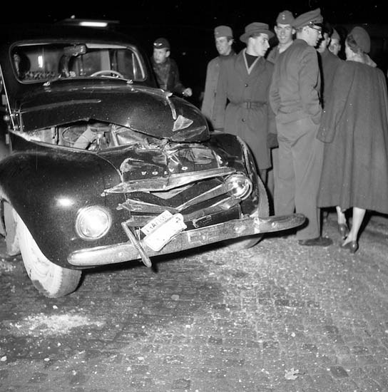 Schwarz-weißes Bild eines schwer beschädigten Autos am Straßenrand umgeben von einer Gruppe besorgter Menschen.