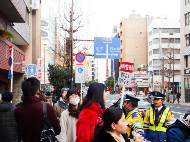 Eine volle Szene mit Menschen in verschiedenen Kleidern und Kappen, einer Handtasche, Haarsträhnen, Brett, Pfahl, Bäume, Gebäude mit Fenstern, eine Straße mit einer weißen Linie und einem weißen Himmel.