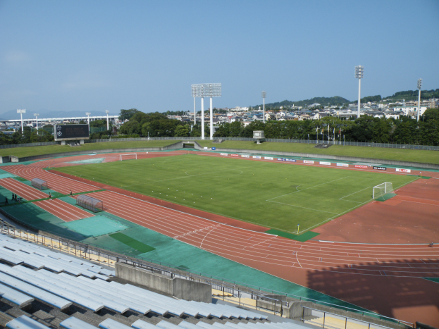 Ein großes Stadion mit einem Fußballfeld, umgeben von grünem Gras, Bäumen und Laternen, mit ein paar Menschen auf dem Boden, leeren Stühlen, fernen Gebäuden und einem klaren blauen Himmel.