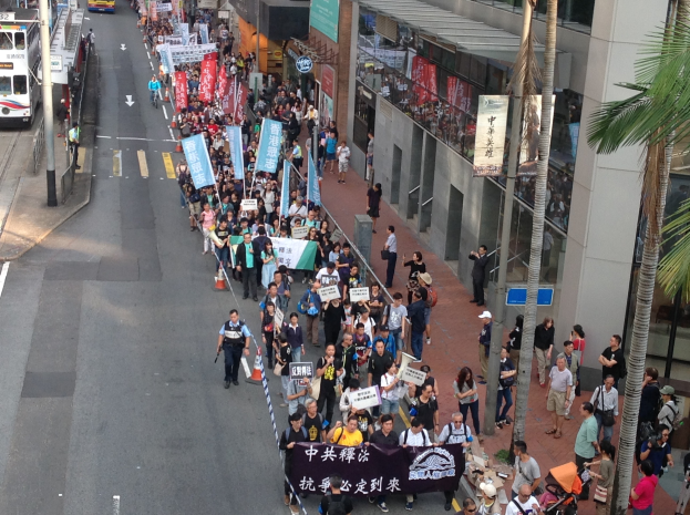 Große Menschenmenge marschiert bei einer Demonstration auf einer Stadtstraße, hält Schilder und Plakate hoch, mit Bäumen, Glasgebäuden und Fahrzeugen im Hintergrund.