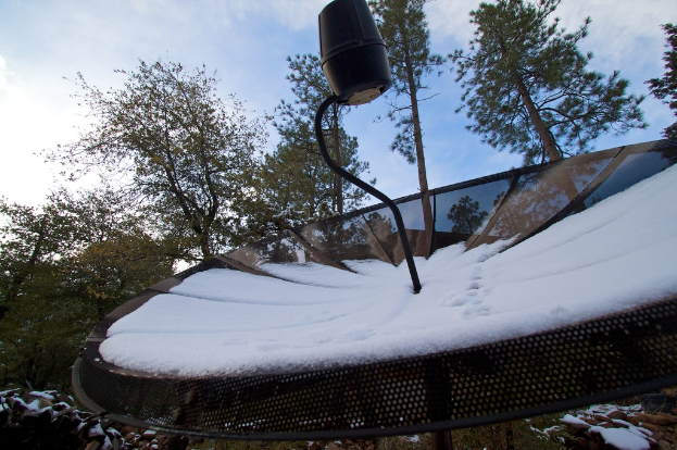 Eine Schüsselantenne mit Schnee darauf, vor einem Hintergrund von ein paar Bäumen unter einem klaren Himmel.
