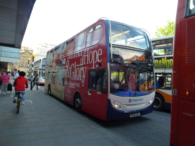 Eine Person fährt mit dem Fahrrad auf einem Bürgersteig mit vielen Menschen, Bussen mit Text, Gebäuden und Laternen im Hintergrund.