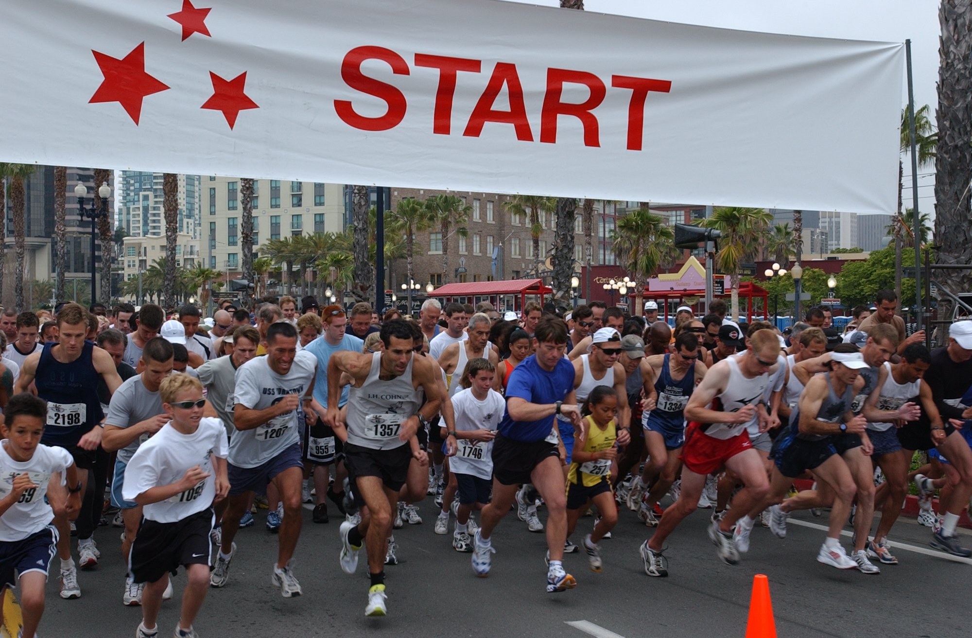 Gruppe von Menschen, die bei einem Marathon laufen, mit einem Verkehrskegel im Vordergrund und einer Fahne im Hintergrund, umgeben von Bäumen, Laternenmasten, Gebäuden und einem klaren blauen Himmel.