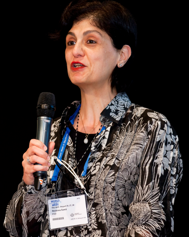 Eine Frau in einem schwarzen Hemd mit einem weißen Design, die ein blaues Abzeichen trägt, hält ein Mikrofon und spricht.