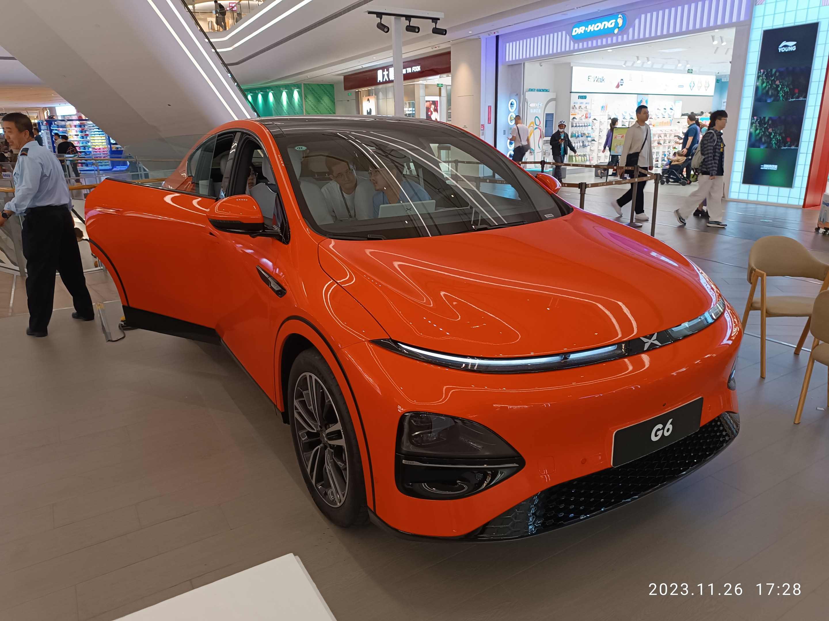 Orangefarbener Elektroauto in einer Ausstellungshalle umgeben von Menschen, Stühlen, einem Wagen, Geschäften und Namensschildern im Hintergrund.