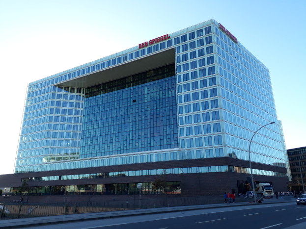 Ein großes Gebäude mit vielen Fenstern, das als Hauptquartier der Deutschen Bank identifiziert wurde, umgeben von Straßenmasten, Straßenlaternen, Kraftfahrzeugen, Fußgängern, Bäumen und einem bewölkten Himmel.