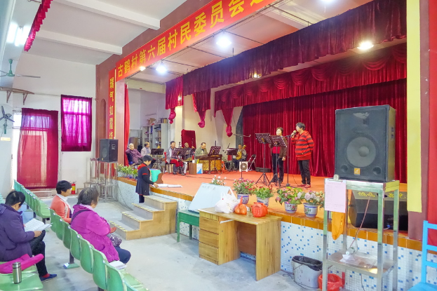 Eine Bühne mit zwei singenden Personen, einer Band im Hintergrund, roten Vorhängen, einer Lampe, einem Lautsprecher rechts und einem Publikum auf Stühlen sitzend.