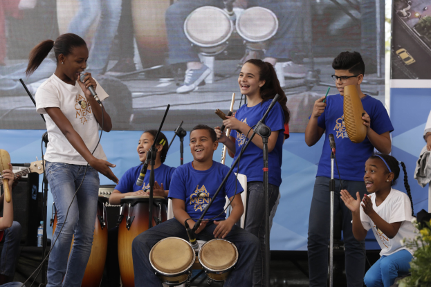Kinder auf der Bühne mit Musikinstrumenten, eine Frau mit Mikrofon, Mikrofonständer, ein Banner im Hintergrund und Topfpflanzen rechts.