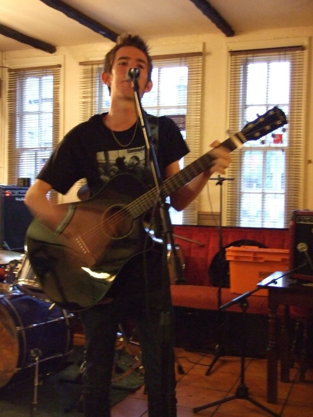 Ein Mann in schwarzem T-Shirt und Hose steht mit einer Gitarre und einem Mikrofon auf einem Ständer, mit einer blauen Trommel in der Ecke, einer Couch mit Gegenständen darauf und durchsichtigen Vorhängen im Hintergrund.