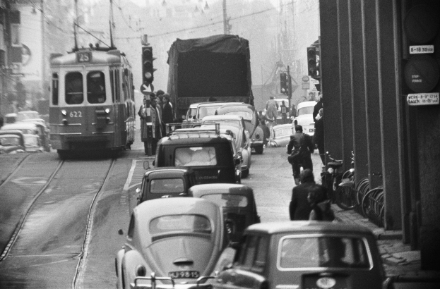 Ein Schwarz-Weiß-Foto einer belebten Stadtstraße mit zahlreichen Autos, Lastwagen, Fußgängern, parkenden Fahrrädern und Gebäuden, mit Verkehrssignalen in der Ferne.