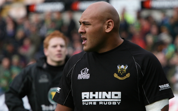 Zwei Männer stehen, einer schaut nach links und scheint zu sprechen, mit einer unscharfen Menge im Hintergrund.