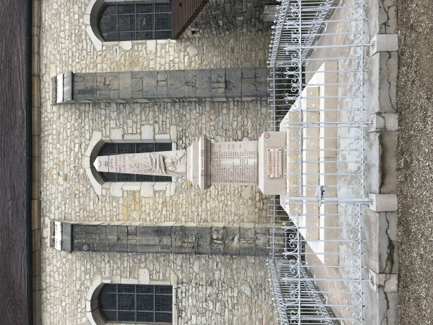 Steinbau mit Metallzaun, einer Säule mit Text und Fenstern, neben einer Straße gelegen.