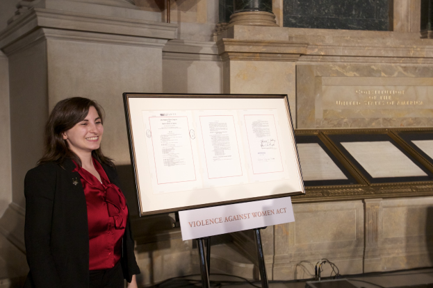 Eine Frau in einem roten Hemd und schwarzer Jacke lächelt vor einem Schild, auf dem "Gesetz gegen Gewalt an Frauen" steht, mit einer Wand und Säulen im Hintergrund.