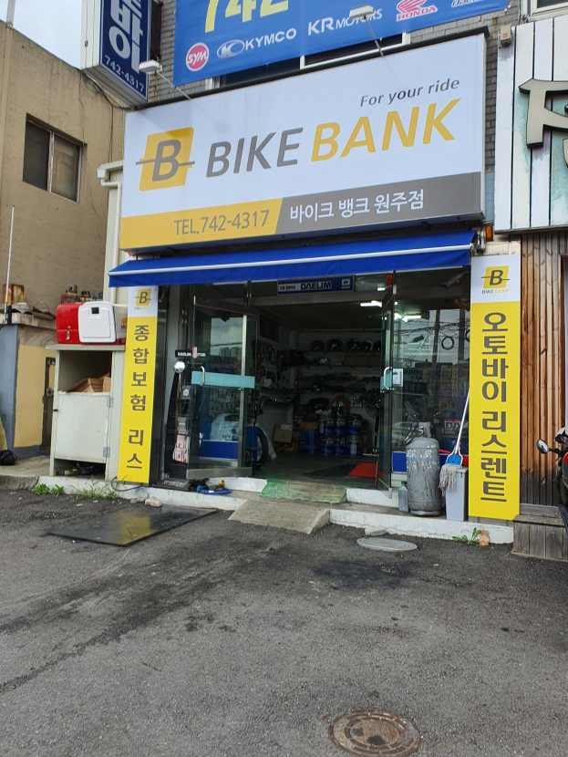 Außenansicht eines Fahrradgeschäfts mit einem "Bike Bank"-Schild, parkende Fahrzeuge davor, eine Straße rechts daneben und sichtbare Texttafeln im Innern.