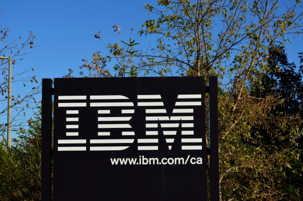 Das IBM-Logo vor dem Firmensitz in San Francisco, Kalifornien, mit einer schwarzen Tafel im Vordergrund, auf der der Text 'IBM' steht, und einigen Bäumen im Hintergrund, die von einer Straßenlaterne beleuchtet werden und vor einem klaren blauen Himmel stehen.