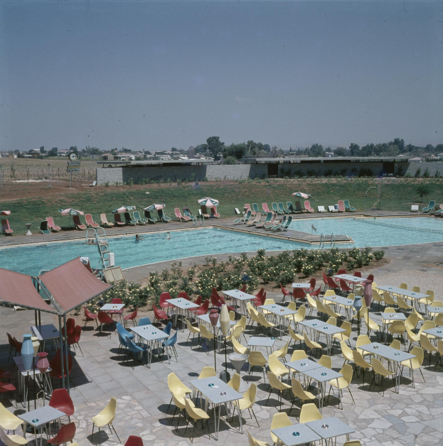 Freibad mit Tischen, Stühlen, Pflanzen, Sonnenschirmen und umgebenen Strukturen unter einem bewölkten Himmel.