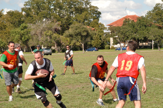 Menschen spielen American Football auf einem grünen Feld mit einem Haus, Autos und Bäumen im Hintergrund.