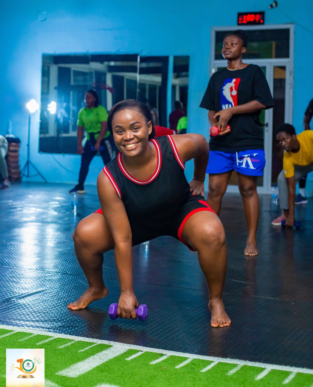 Eine Frau in einer Kniebeugeposition, die Hanteln in einem Fitnessstudio mit Equipment und Branding im Hintergrund hält.