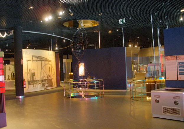 Großer Raum mit verschiedenen Ausrüstungsgegenständen, einschließlich eines Metallobjekts auf dem Boden, einer Säule, einem Feuerlöscher, Texttafeln, Metallstangen, einem Schild und einer Vitrine.