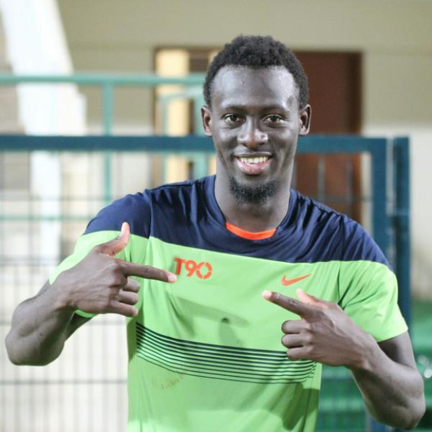 Ein Mann in einem grünen und blauen Hemd, identifiziert als Emmanuel Adebayor, zeigt mit dem Finger auf die Kamera und lächelt, mit einem unscharfen Geländer, Wand und Tür im Hintergrund.