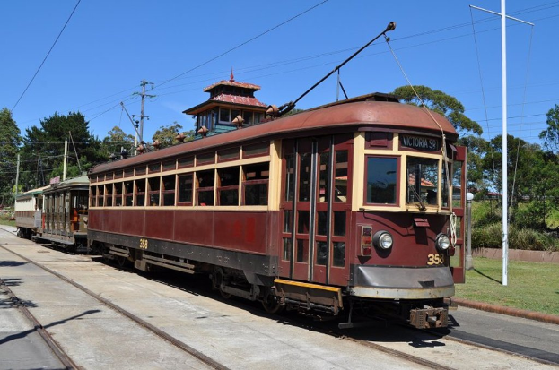 Ein Zug auf Schienen mit Pfählen, Kabeln, Bäumen und Häusern im Hintergrund.