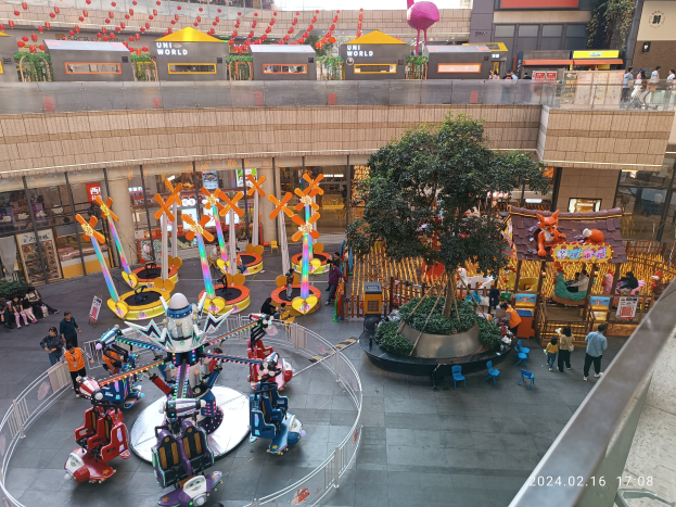 Ein Vergnügungspark in einem Einkaufszentrum mit verschiedenen Attraktionen, Bäumen, Menschen, Stühlen, Ständen und Gebäuden sowie einem Wasserzeichen unten rechts.