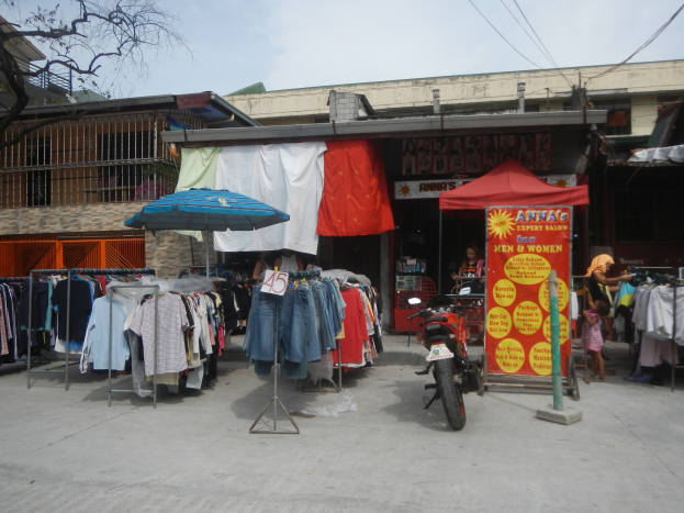 Eine Straßenszene mit einem vor einem Bekleidungsgeschäft geparkten Motorrad, das Kleidungsstücke auf Bügeln, einen Schirm und eine Person in der Nähe zeigt, mit Gebäuden, einem Baum und dem Himmel im Hintergrund.
