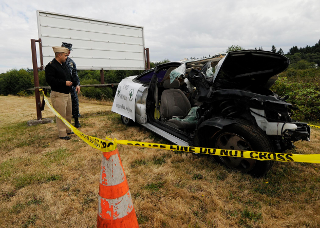 Ein Polizist in Uniform steht neben einem schwer beschädigten Auto am Straßenrand, mit einem Verkehrskegel und einem Band an einem Pfahl in der Nähe und Bäumen und einem bewölkten Himmel im Hintergrund.