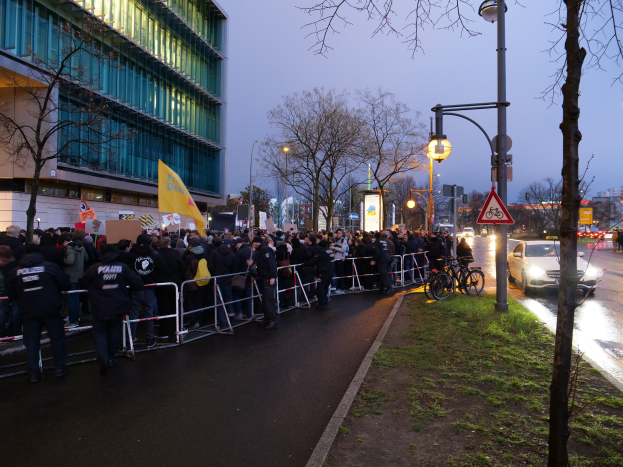 Große Menschenmenge protestiert vor einem Gebäude in Berlin, hält Schilder, mit Barrikaden, Fahrrädern, Laternenmasten, Schildern, Bäumen und Gras im Hintergrund.
