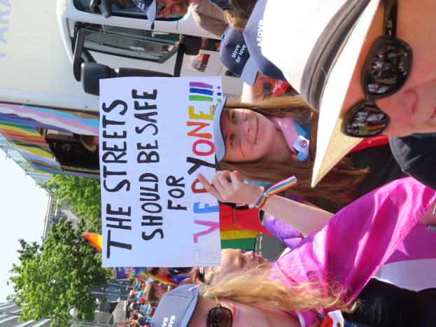 Eine Gruppe von Menschen hält ein Schild mit der Aufschrift "Die Straßen sollten sicher sein" vor einem Lastwagen, einige tragen Mützen und Schutzbrillen, andere halten Schilder, vor einem Hintergrund aus Bäumen, Gebäuden und einem klaren blauen Himmel.