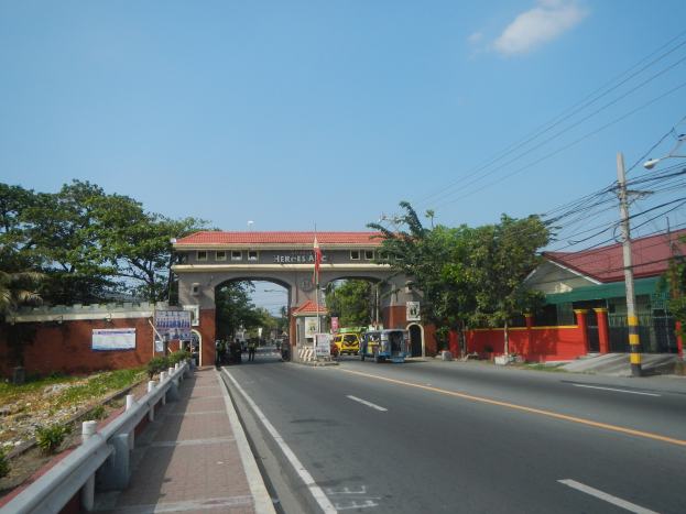Eine Straße mit fahrenden Autos, ein Fußweg mit Geländern auf der linken Seite, Bäume und Gebäude auf beiden Seiten, ein Tor mit einer Namensschild, Strommasten mit Drähten und ein bewölkter Himmel im Hintergrund.