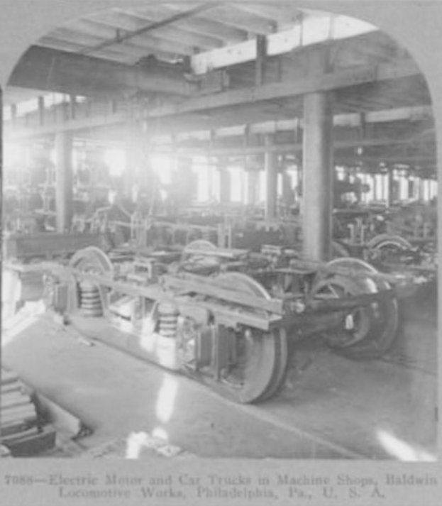 Schwarzes und weißes Foto eines Elektromotors und eines Lkw in einer Fabrik mit Maschinen, Säulen und einem Dach, mit Text am unteren Rand.