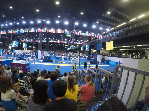 Eine große Menschenmenge sitzt und steht in einer Indoor-Sportarena mit Bildschirmen, Flaggen und Deckenbeleuchtung im Hintergrund.