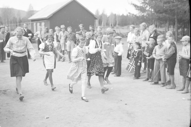 Schwarzes Bild von Kindern, die vor einer Menge mit Fahnen Fußball spielen, mit Bäumen und einem Haus im Hintergrund.
