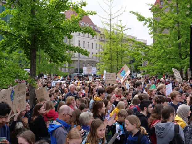 Eine große Menschenmenge protestiert vor einem Gebäude in Berlin, hält Schilder hoch, mit Bäumen, Fahrzeugen, einem Lautsprecher und dem Himmel im Hintergrund.