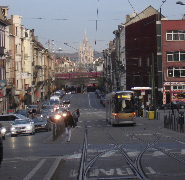 Eine belebte Stadtstraße mit Fahrzeugen, Fußgängern, einer Tram, Gebäuden, einer Brücke, Bäumen und einem klaren blauen Himmel.