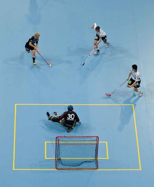 Drei Personen in Sportbekleidung spielen Hockey, wobei eine Person rechts mit einem Hockeystock einen Ball schlägt und eine Person hinter einem Tor auf dem Boden sitzt.