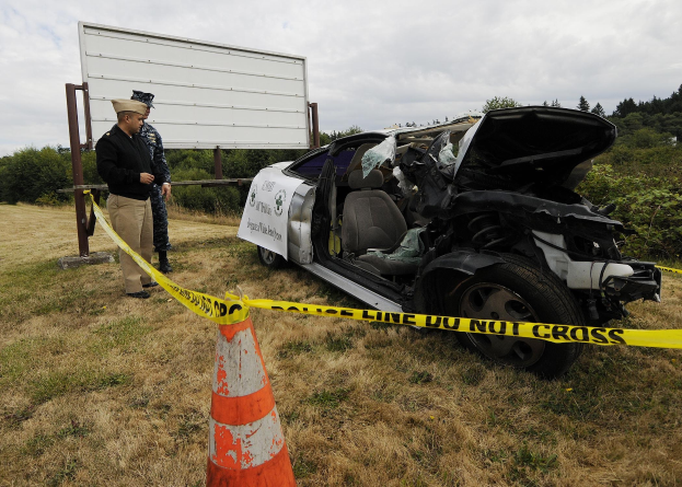 Polizeibeamter in Uniform inspiziert ein schwer beschädigtes Auto mit eingedellter Haube und zersplitterten Fenstern am Straßenrand, mit einem Verkehrskegel und Band in der Nähe vor dem Hintergrund von Bäumen und einem bewölkten Himmel.