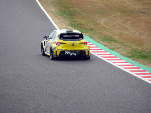 Ein gelbes Sportauto mit Text und Zahlen darauf, das eine Rennstrecke entlangfährt, umgeben von Gras.