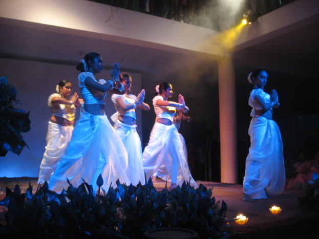 Eine Gruppe von Menschen tanzt auf der Bühne mit einer Gruppe von Pflanzen vor ihnen, während eine andere Gruppe von Menschen zusieht.