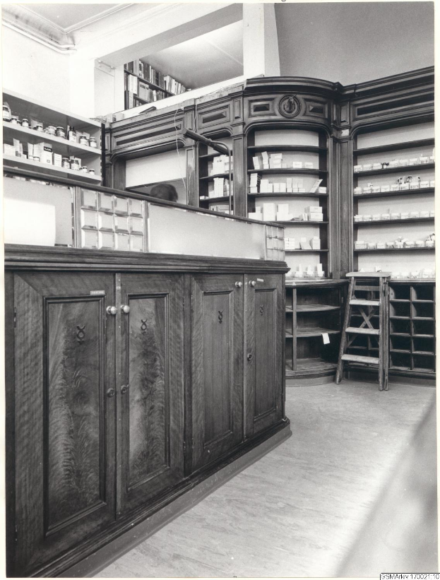 Ein Schwarz-Weiß-Foto einer Apothekeninnenseite mit einem Tresen, Schränken, einer Leiter und Regalen, die mit verschiedenen Gegenständen gefüllt sind, mit Text oben.