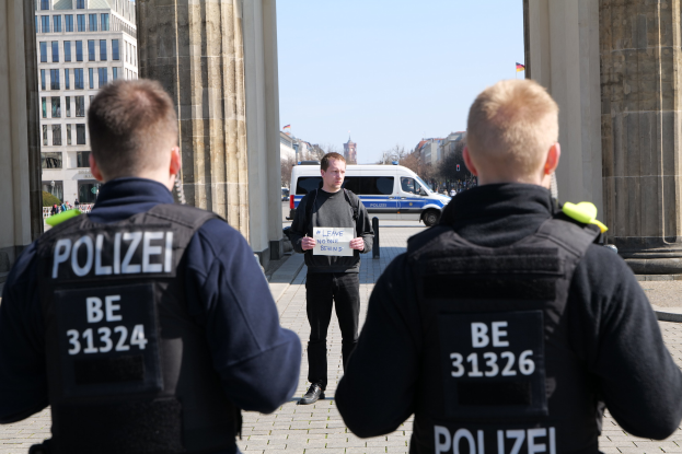 Drei Polizeibeamte stehen vor einem Gebäude, wobei ein Mann in der Mitte ein Papier hält, flankiert von Säulen und einem belebten städtischen Hintergrund.