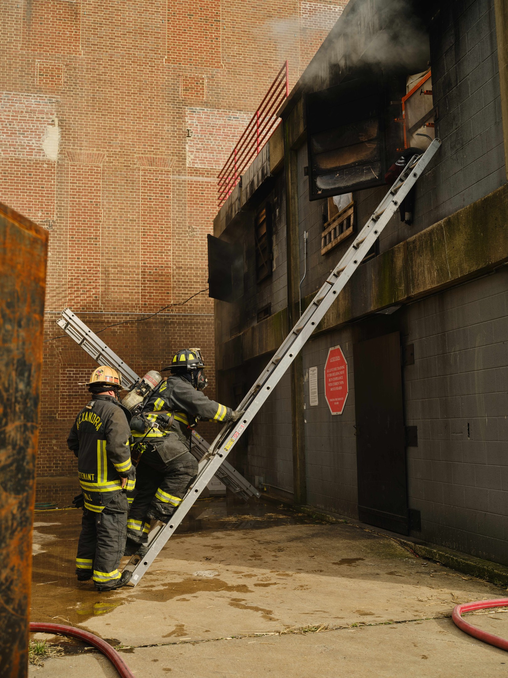Feuerwehrleute in Helmen klettern an einer Leiter zu einem von Rauch erfüllten Gebäude hoch, mit einem Metallobjekt links und sichtbaren Rohren am Boden.