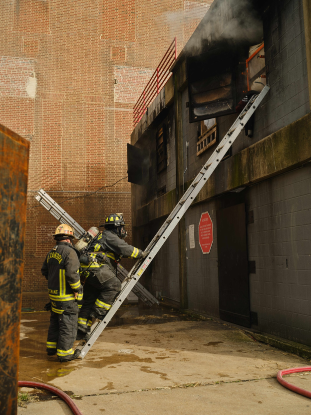 Feuerwehrleute in Helmen klettern an einer Leiter zu einem von Rauch erfüllten Gebäude hoch, mit einem Metallobjekt links und sichtbaren Rohren am Boden.