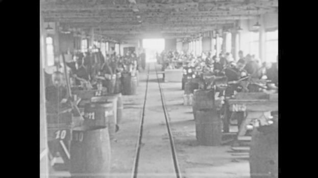 Schwarzes und weißes Foto einer Fabrik mit Arbeitern, die Maschinen bedienen, nummerierten Fässern, einer Eisenbahnschiene, Säulen und Deckenlampen, die die industrielle Revolution widerspiegeln.