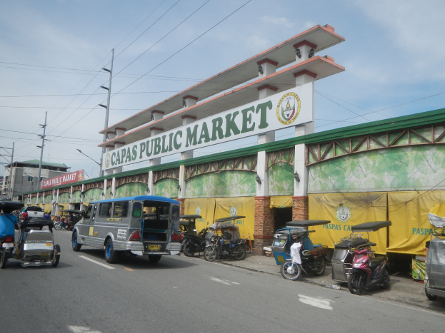 Eine belebte Stadtstraße mit Autos, Motorrädern und Rikschas vor einem Gebäude mit der Aufschrift "Capas Public Market", mit Strommasten, Laternenmasten und bewölktem Himmel im Hintergrund.