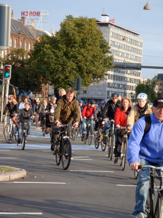 Menschen, die auf einer Straße mit Pfosten, Bäumen, Gebäuden und dem Himmel im Hintergrund Fahrrad fahren.