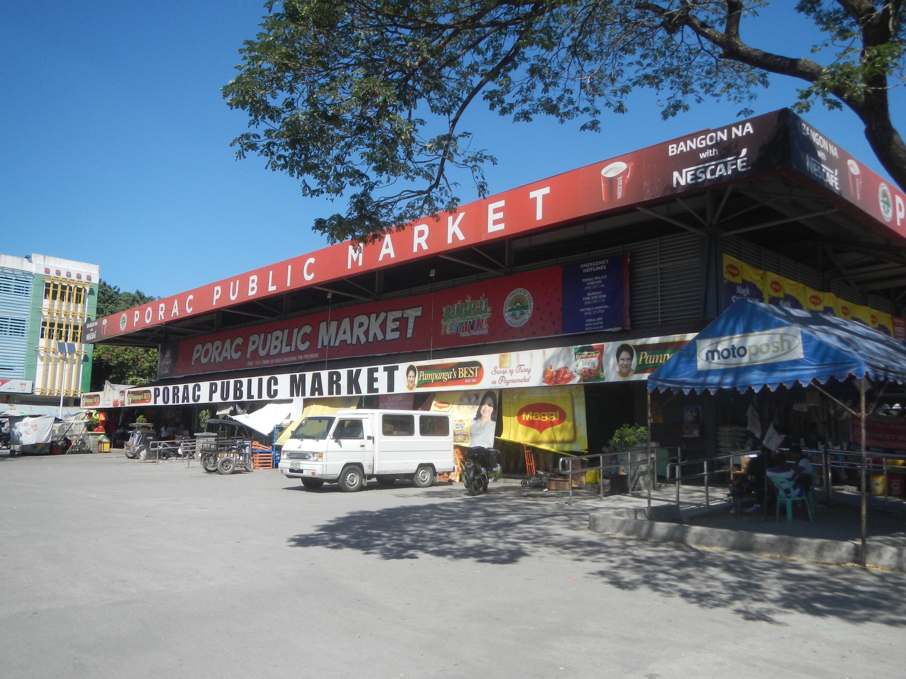 Öffentlicher Markt mit einem vor ihm geparkten Van, umgeben von Gebäuden, Motorfahrzeugen, Personen unter einem Zelt, Geländern, Bäumen und einem klaren blauen Himmel.